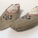 Image shows a pair of grey buckskin moccasins with colourful embroidered patterns and a red trim.