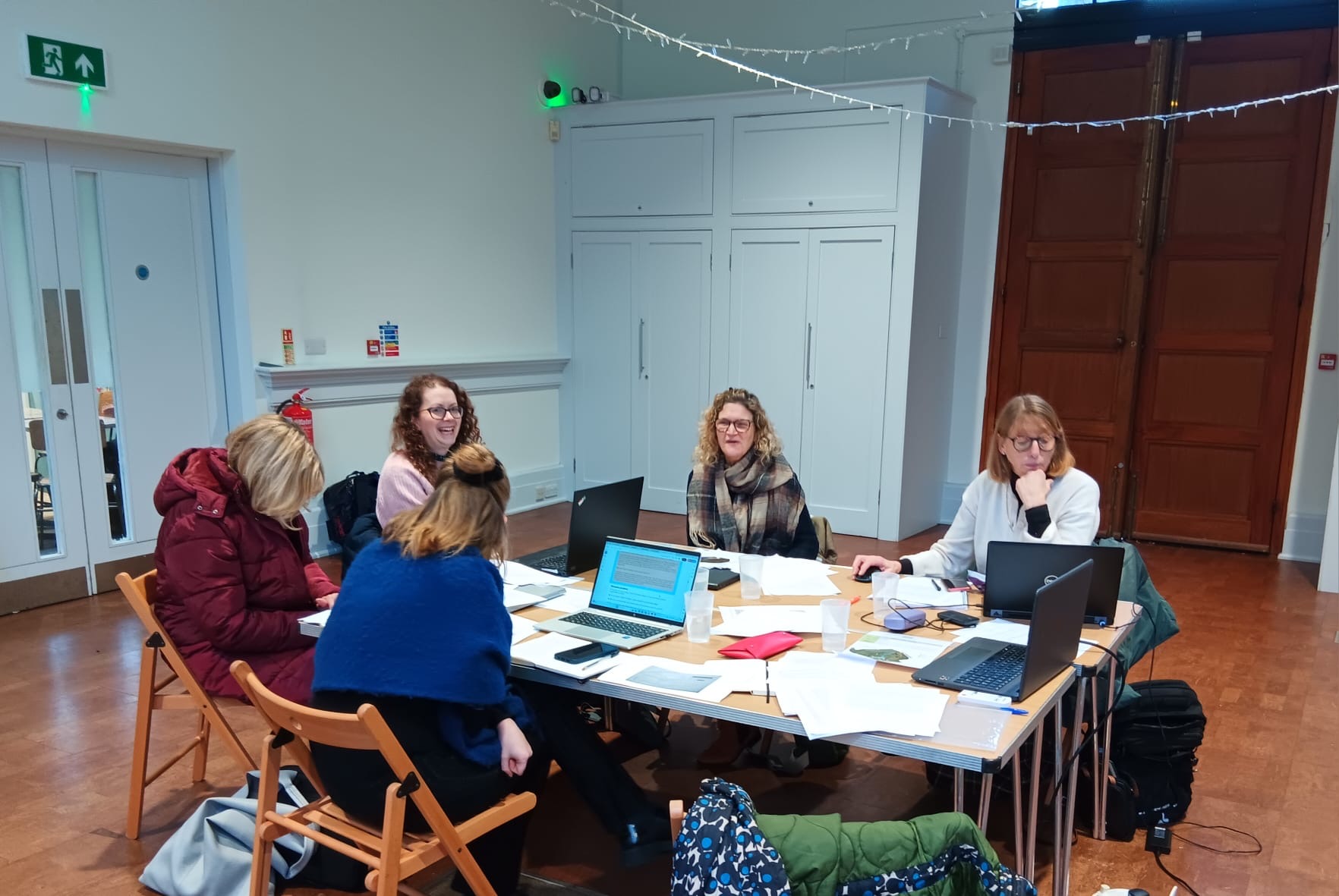 Image shows a group of women sitting in a room around a table. On the table, you can see papers and laptops.
