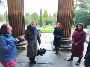 Image shows a group of women. One woman, wearing a blue coat, is talking. The others are looking and her and listening. They are stood in front of two large pillars.
