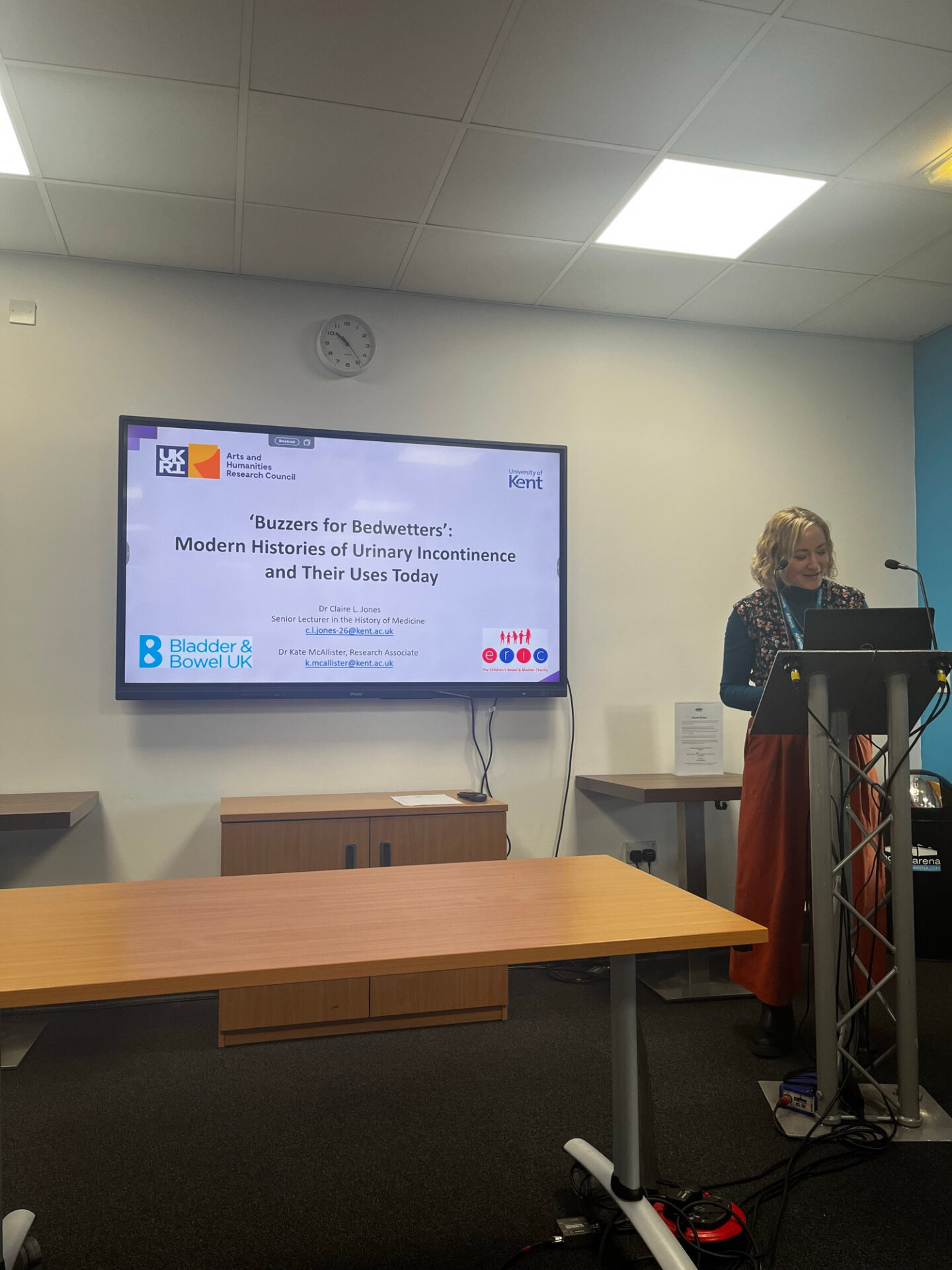 Woman standing behind a lecturn, looking down at her notes. Behind her, there is a screen showing an introductory PowerPoint slide.