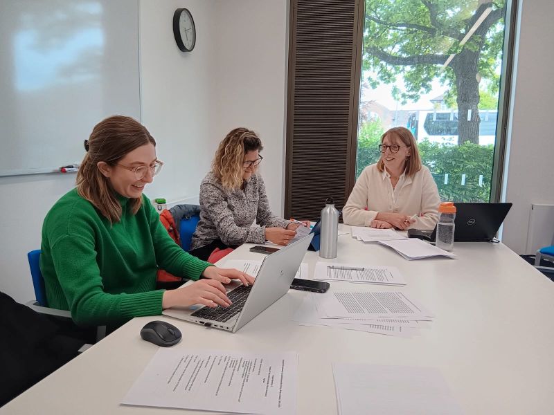 Image shows three women sat around a table. Two of the women are smiling and looking down, whilst the other is looking at the others.