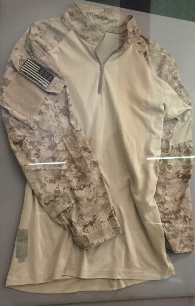 Photograph of a camouflage jacket in a clear glass case. There is an inverted U.S. flag on the right sleeve, which is displayed to face the viewer. 