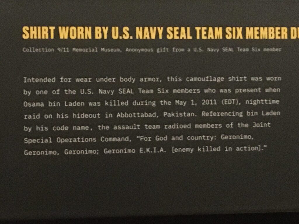 Photograph of a museum label taken in the 9/11 Memorial & Museum. The caption reads: 'Intended for wear under body armor, this camouflage shirt was worn by one of the U.S. Navy SEAL Team Six members who was present when Osama bin Laden was killed during the May 1, 2011 (EDT), nighttime raid on his hideout in Abbottabad, Pakistan. Referencing bin Laden by his codename, the assault team radioed members of the Joint Special Operations Command, "For God and country: Geronimo, Geronimo, Geronimo; Geronimo E.K.I.A. (enemy killed in action)."'