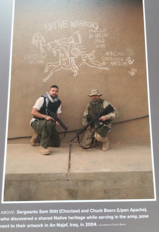 Photograph of two U.S. soldiers posing in front of a graffitied wall. One wears a black and white scarf around his neck and the other wears a camouflage hat. Both are holding their guns. The graffiti depicts a man on horseback wearing a feathered headdress. The words read: 'NATIVE WARRIORS, Al Najaf, Iraq, 2004' and the names of the Choctaw, Apache and Cherokee Nations.