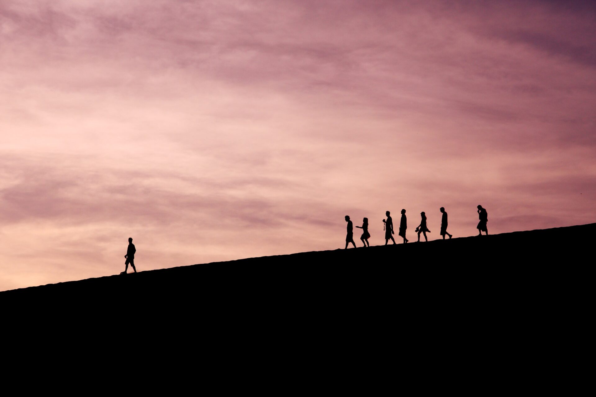 Silhouettes of seven people walking in a line downhill, following a single lead person.